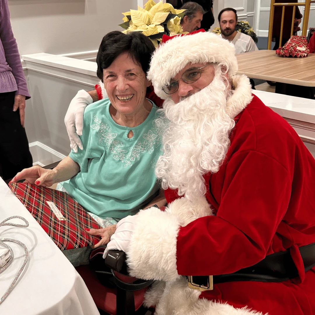Smiling Mount Sinai resident sits with Santa holding a wrapped gift