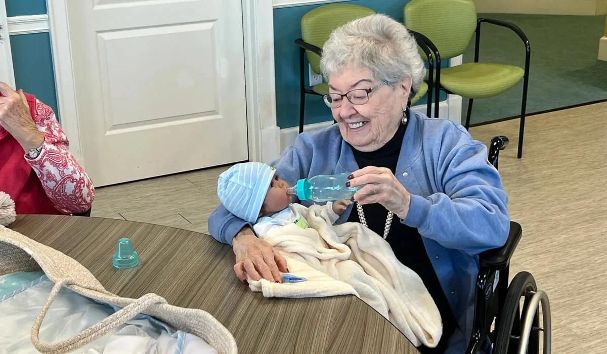 Resident holds a baby doll and pretends to feed it from a bottle while sharing stories and smiling